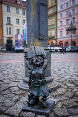 Polonya 'nın Wroclaw kentinde bir sokak lambasının yanında açık bir kitap tutan bronz bir cüce. Pastel konaklarının arkası bulanık, yumuşak bulutlu gün ışığı altında sığ alan derinliği.