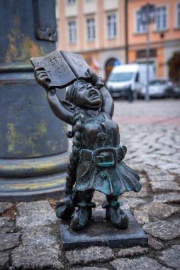Bronz cüce heykeli, Polonya 'nın Wroclaw Eski Kasabası' ndaki bir kaldırım taşı caddesinde açık bir kitabı kaldırıyor. Pastel konutlar ve bir sokak lambası bulutlu bir günde arkasında bulanıklaşır.
