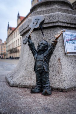 Bronz bir cüce, Polonya 'nın Wroclaw şehrinde taş bir kaideyle kollarını kaldırmış bir gitar tutuyor. Tarihsel cepheler ve kırmızı kiremitli çatılar gündüz vakti bulutlu ışıkta bulanıklaşır.