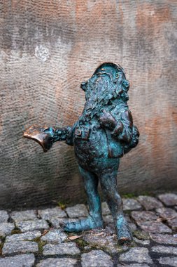 Bronz Kıskaç cüce, yıpranmış bir duvarın yanında kaldırım taşlarının üzerinde duruyor. Sırt çantası ve kamerasıyla açık bir kitap tutuyor. Yumuşak gün ışığında göz düzeyi metal parçacıkları gösteriyor.