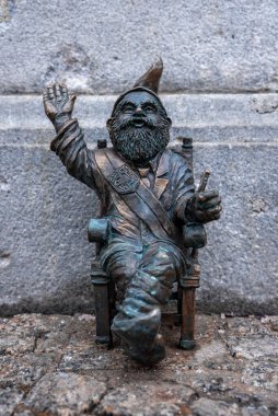 Bronz bir Wroclaw Krasnale cücesi ahşap bir sandalyede oturur, dalgalar, ve sert bir taş duvarla kurulmuş bir asa tutar ve gün ışığında yumuşak doğal ışıkla kaldırım taşlarını taşır..