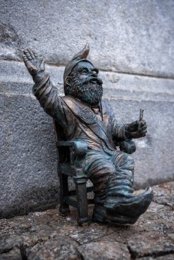 Wroclaw, Polonya bronz cücesi sivri uçlu şapkalı bir sandalyede taş bir duvarın yanında oturuyor, bir kolu havada, diğeri küçük bir anahtar ya da alet tutuyor, zemin seviyesinde yumuşak bir ışık.