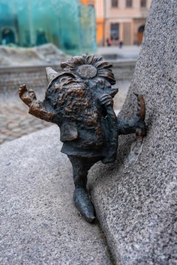 Bronz bir Wroclaw cücesi, Polonya 'nın Wroclaw kentindeki Plac Golebi' nin cam çeşmesinin yanındaki bir ayçiçeğiyle kaya çıkıntısına tırmanır. Yumuşak gün ışığı ve sığ odak ayrıntıları gösterir.