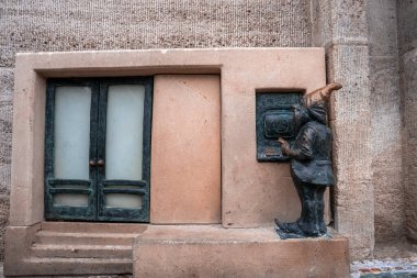 Polonya 'nın Wroclaw kentindeki minyatür bir ATM' de bronz bir cüce duruyor. Gün ışığı desenli taşı, sessiz tonları ve şehir duvarındaki küçük çift kapılı şakacı ölçeği ortaya çıkarır..