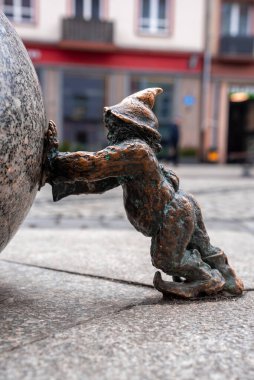 Wroclaw, Polonya 'da bronz bir cüce taş kaldırımlı bir caddede granit bir küre itiyor, zemin seviyesi, gün ışığı, yüzeysel alan derinliği, yan profil bileşimi.