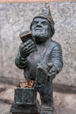 Yakından bakınca, Wroclaw cüce bronz heykeli taş bir duvarın yanında, bir levha tutuyor ve bozuk para kutusunun üzerinden bir çanta açıyor, sivri uçlu şapka ve sakal gün ışığında parlıyor.