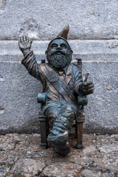 Bronz bir Wroclaw Krasnale cücesi ahşap bir sandalyede oturur, dalgalar, ve sert bir taş duvarla kurulmuş bir asa tutar ve gün ışığında yumuşak doğal ışıkla kaldırım taşlarını taşır..