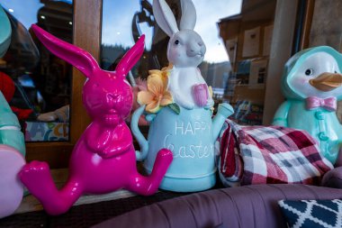 Bir vitrin vitrininde seksi pembe bir tavşan, pastel tavşan sulama kutusu Mutlu Paskalyalar, kumaş çiçekler, pembe lale, ekose fırlatma ve bir deniz ördeği görülüyor..