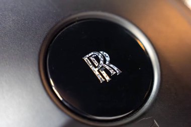 Sıkı bir yakın çekim, krom RR monogramı ile parlak siyah direksiyon merkezini çerçeveler. Sığ alan derinliği ve sıcak gölgeler stüdyo ışıklandırması ve lüks.
