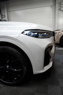 Ön sol BMW X7, yontulmuş tampon, mavi aksanlı lazer far, yuvarlak tekerlekli siyah tekerlek, park sensörü, parlatıcı aero girişleri, endüstriyel duvarlar ve lastik zemin.