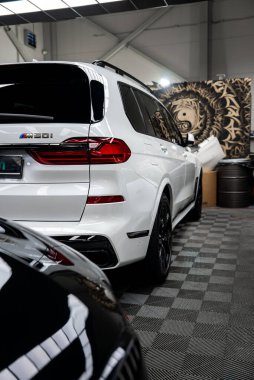 Beyaz BMW X7 M50i kapalı bir tamirhanede oturuyor. Kareli zemini, endüstriyel ışıkları, istiflenmiş tekerlekleri, parlak siyah jantları, LED arka lambaları ve yontulmuş tamponu var..