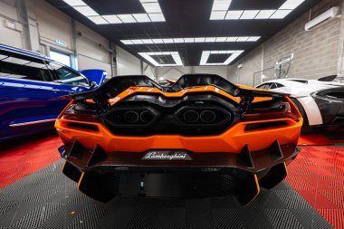 Turuncu Lamborghini Revuelto modern bir atölyede kapalı duruyor, avluda yüksek altıgen egzozlar, karbon dağıtıcı yüzgeçler, açısal LED stop lambaları, solda mavi araba, sağda beyaz süper araba..