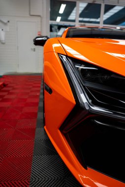 Turuncu Lamborghini Revuelto 'nun sol ön köşesinde bir atölyede, Jilet LED far, açısal giriş, siyah aero, kırmızı ve siyah fayanslar, ve sığ alan derinliği.
