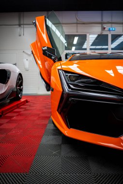 Turuncu Lamborghini Revuelto 'nun sağ ön çeyreğinde yükseltilmiş makas kapısı, Y şeklinde LED far, karbon fiber emme, ışık yansımaları, fayanslar, asansör, servis bölmesi.