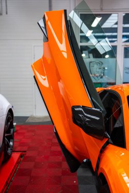 Turuncu Lamborghini Revuelto kapısı, kırmızı kareli fayanslar, parlak ışıklandırma, karbon fiber ayna ve soldaki asansörde bir başka performans arabası ile bir atölyede yükselir..
