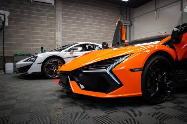 Canlı turuncu bir Lamborghini Revuelto kapalı bir garajda makas kapısı açık, açılı LED farlar, parlak siyah tekerlekler ve yanında park etmiş beyaz bir McLaren ile oturuyor..