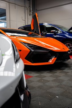 Turuncu Lamborghini Revuelto bir servis hangarında oturuyor, makas kapısı açık, LED farlar ve altın boğa rozeti görünür, mavi hatchback ve gri spor arabası da yakında..