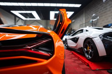 Turuncu Lamborghini Revuelto arkadan Z şeklinde stop lambası ve modern bir atölyede makas kapısı, kırmızı kauçuk zemin, şebeke LED ışıkları, beton duvarlar, asansördeki McLaren..