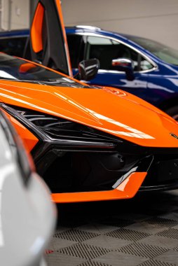 Turuncu Lamborghini Revuelto 'nun sol ön çeyreği bir atölyede, makas kapısı kaldırılmış, Y şeklinde LED far ve hava emişi görünür, parlak kaput yansıtıcısı dükkan ışıkları.