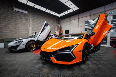Makas kapıları ve LED Y farları olan turuncu Lamborghini Revuelto karbon aksanlı ve desenli zemin desenli parlak bir panel aydınlatma atölyesinde beyaz McLaren 'ın yanında oturur.
