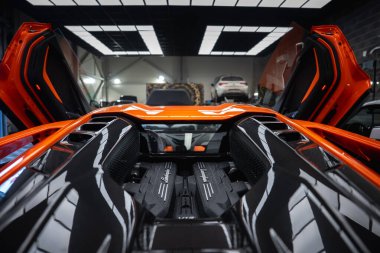 Turuncu Lamborghini Revuelto makas kapıları açık bir atölyede duruyor, V12 motor bankaları Lamborghini betiği, karbon fiber paneller, araçlar ve görünürde asansörler gösteriyor.