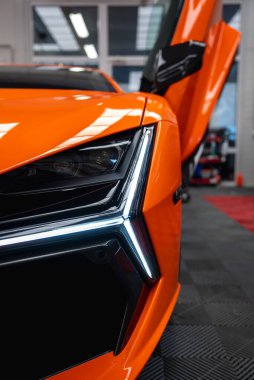 Bir atölyede turuncu bir Lamborghini Revuelto 'nun yakın görüntüsü, sol ön köşede, Y şeklinde LED far, keskin fasya, yükseltilmiş makas kapısı, alet rafları, lastik fayanslar, floresan lambalar..