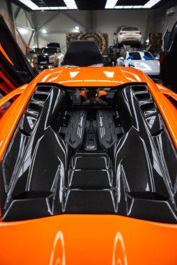 Turuncu bir Lamborghini Revuelto arkadan monte edilmiş siyah kapaklı ve havalandırmalı V12 'sini bir servis garajında makas kapıları ve aletler ve asansörlerle gösteriyor..