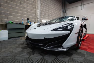Bir McLaren P1 atölyesinde oturur, çerçeveye doğru burun, bumerang LED farlar, karbon ayırıcı, oyulmuş kaput kapakları, sarı kalibreler, parlak endüstriyel aydınlatma, kırmızı ve siyah fayanslar.