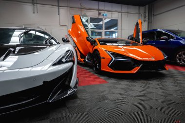 Kapalı atölye sahnesi, gümüş renkli bir McLaren P1 'in turuncu bir Lamborghini' ye baktığını gösteriyor. Her ikisi de siyah ve kırmızı kareli bir zeminde parlak ışık altında yükseltilmiş makas kapılı..