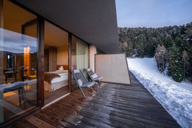 Çağdaş bir otel odası Dolomitler 'de ahşap bir terasa açılıyor. İki şezlong karlı bir yamaçla ve sonsuz yeşil bir ormanla yüzleşir. Alacakaranlık ışığı sıcak içiyle buluşur..