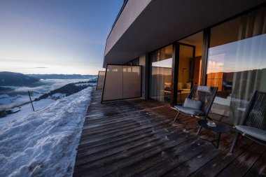 Dolomitlerdeki modern süit terası alacakaranlıkta karlı vadiye açılıyor. Alçak bulutlar tepeler arasında dinleniyor, pastel gökyüzü soluyor, sıcak iç ışık cam kapılara yansıyor..