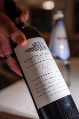 Tenuta di Arceno Chianti Classico Riserva 2020, Tuscan Cypress tepeleri ve çiftlik evi ile krema etiketli, Dolomitlerde sıcak bir yemek odasında.
