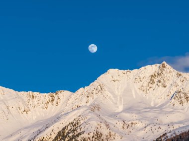 Dolunay, kuzey İtalya 'daki sivri uçlu bir Dolomite zirvesinin üzerinde. Kar dolu zirveler kobalt gökyüzünün altında gün batımında ışıldar, uzun bir mercekle çekilir..