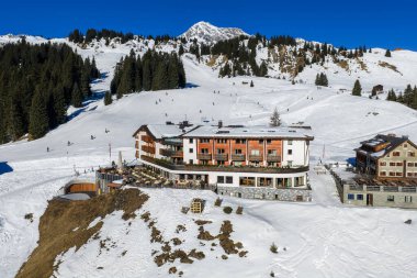 Lech ve Oberlech 'teki bakımlı pistlerin yanında lüks bir otelin yüksek açılı kış manzarası. Kayakçılarla güneşli yamaçlar, koyu yeşillikler ve gün ortası ışığı altında sivri bir kar zirvesi..