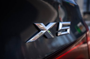 Yakından bakınca parlak siyah bir BMW 'nin arkasındaki krom X5 rozeti görünüyor. Yumuşak gün ışığı parlak yüzeyleri yansıtıyor. Bulanık kentsel formlar arkada oturuyor..