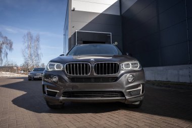 BMW X5 'e bakan ön cephede böbrek ızgaraları, LED korona halka farları ve HO 3232 plakalı koyu renk ağaçlarla kaplı bir sanayi binası var..