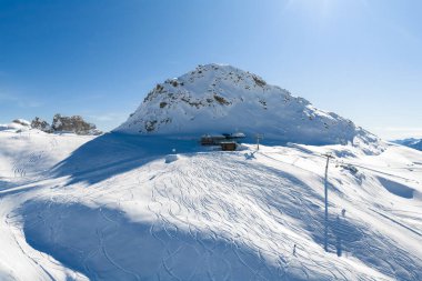 İtalyan Alplerindeki San Domenico kayak merkezinin hava manzarası kayalık bir tepe, kayak kaldırma istasyonu ve sütunlar, bakımlı pistler, taze izler ve yamaçlardaki küçük figürler gösteriyor..