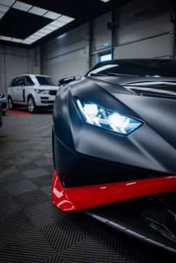 Matte siyah Lamborghini Huracan, kırmızı halı şeridi üzerinde modern SUV ile iyi aydınlatılmış bir galeride LED far ve kırmızı ayracı gösteriyor..