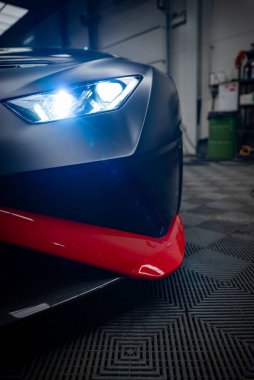 Lamborghini Huracan 'ın bir atölyesinin sol ön köşesinde mat siyah gövde, parlak kırmızı ayrıştırıcı, keskin LED far, lastik kiremitler, alet rafları, floresan ışıklandırma görülüyor..