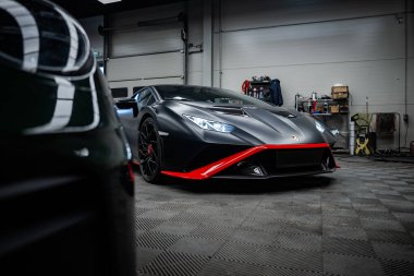 Matte siyah Lamborghini Huracan bir atölyede, LED farlar açık, kırmızı ayırıcı ve yan aksanlar görünür, araçlar ve raflar mevcut, başka bir araba ön tarafta bulanık.