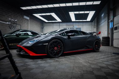 Matte siyah Lamborghini Huracan ayrıntılı bir garajda oturuyor, alçak açılı görüntü aero takımını, kırmızı ayıracı, yan etekleri, arka kanadı, kırmızı kalibreleri, araçları, lastikleri ve ışık panellerini gösteriyor..