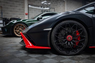 Alçak açı, mat siyah Lamborghini Huracan 'ı kırmızı aksanlı, ayrıştırıcı, orta kilit, kırmızı kalibreli gösteriyor. Yeşil spor araba bir atölyenin arkasında duruyor..