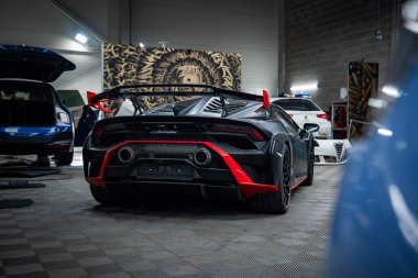 Lamborghini Huracan 'ın arka görüntüsü bir akort stüdyosunda, ayrıntılı ışıklar, açık kapaklar, aslan duvar resmi, karbon kanat, çift egzozlar ve kırmızı süslemelerle çerçevelenmiş bir şekilde oturuyor..