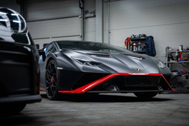 Matte Black Lamborghini Huracan bir tamirhanede oturuyor, önü düşük. Y şeklinde LED farlar, oyulmuş havalandırmalar, siyah tekerlekler, kırmızı kalibreler.