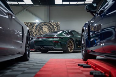 Koyu yeşil Porsche 911, bronz tekerli spor arabaların arasına park edilmiş. Garaj zemininde, dikiz aynasından, çift egzozlu, tavan penceresi aydınlatmalı, asansörlü, aslan duvar resimli..