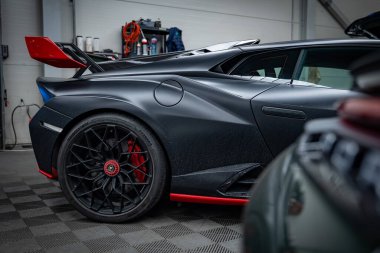Matte Black Lamborghini Huracan bir atölyede sol arka tarafı gösteriyor, alet rafları ve kareli zemin görünür, kırmızı ve siyah arka kanat, dağıtıcı, havalandırma, kırmızı merkez ve kalibreler.