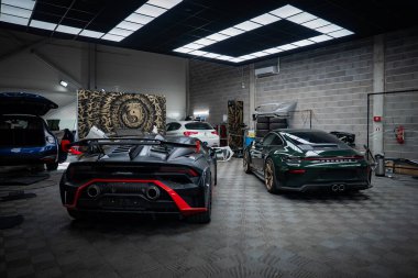 Matte siyah Lamborghini Huracan kırmızı aksanlı arka koltukta oturuyor. Akort garajında, koyu yeşil bir Porsche 911, parlak panel ışıkları, aslan motifli duvar..