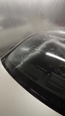 İşçi basıncı araba ön camını köpükle yıkıyor, el kamerasında sarı üniformalı, oto yıkama istasyonunda su jeti kullanıyor..