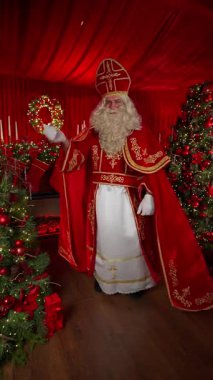 Süslü kırmızı ve beyaz kıyafetli geleneksel Noel figürü süslü şömine ve ağaçların yanında duruyor, altın işlemeli klasik bayram ruhu ve sıcak festival atmosferi..