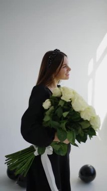 Stüdyoda beyaz güllerle dolu bir buketi olan bir kadın zarif uzun saçlı ve şık siyah elbiseli yumuşak romantik bir ortam yaratıyor..